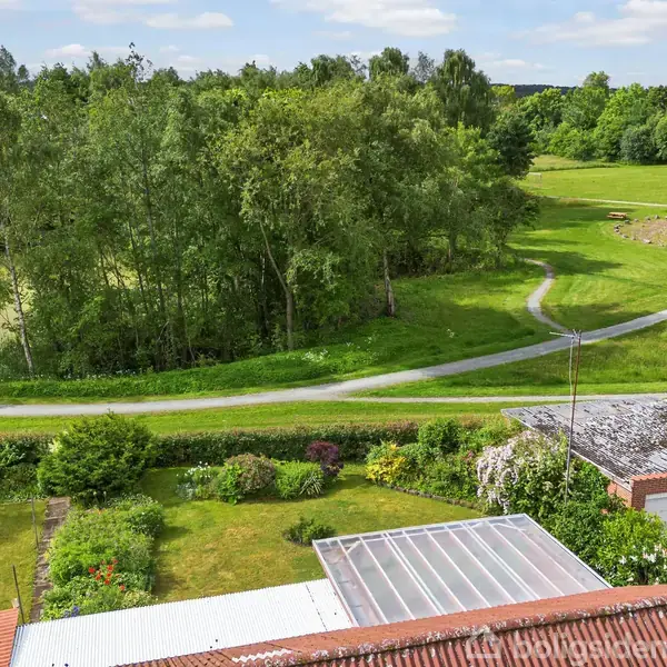 Grøn have med blomster og græsplæne, grænser op til en landevej. Baggrunden viser en park med stier, træer og en klar himmel.