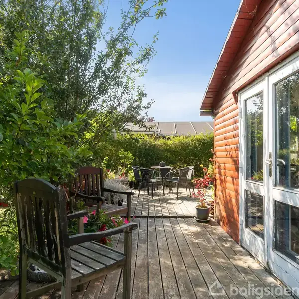 En træterrasse er udstyret med havemøbler og omgivet af frodige grønne planter og buske. Til højre ses en rød træbygning med en stor glasdør.