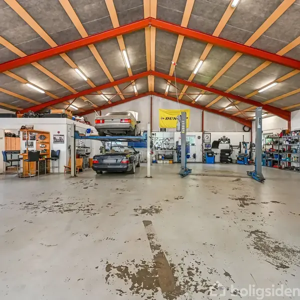 Autoværksted med biler på lifte og værktøj langs væggene. Loftet har røde bjælker og lysrør. På en gul væg hænger et gult skilt med teksten "DEKRA".