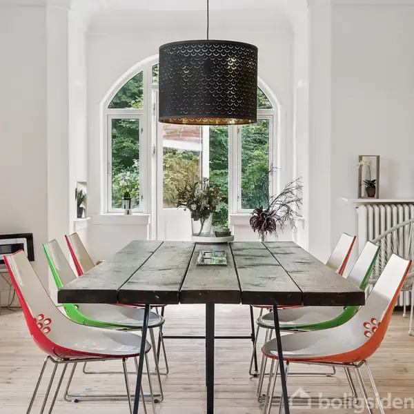 Robust spisebord med træoverflade omgivet af moderne stole i varierede farver. Store vinduer lader naturligt lys ind i rummet, mens en sort pendellampe hænger over bordet. Planter pynter.