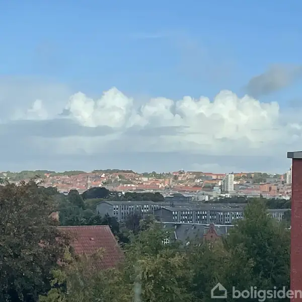 Bygninger og træer fylder forgrunden, mens skyer svæver over en by i baggrunden. En klar blå himmel dominerer ovenfor.