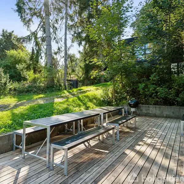 Et metalbord med bænke står stille på en træterrasse ved siden af en grill. Terrassen er omgivet af frodig vegetation og store træer, hvilket skaber en fredelig udendørs atmosfære.