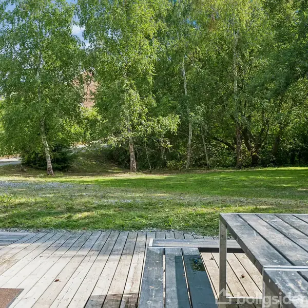 En træterrasse med et bord skaber en afslappende atmosfære, mens grønne træer og en græsplæne udgør baggrunden. Døren til venstre fører ind i huset.
