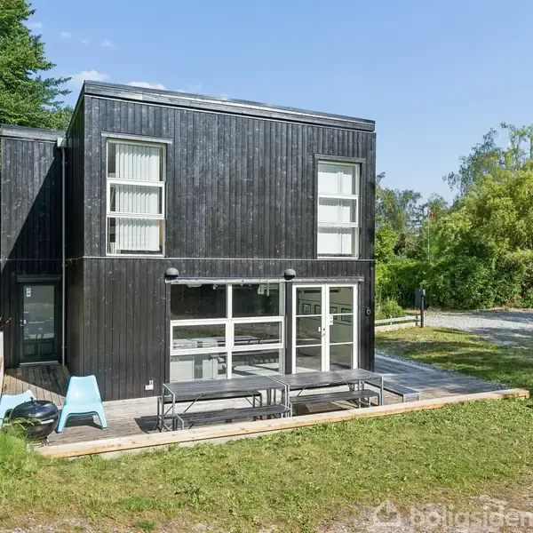 Sort, moderne to-etagers hus med store vinduer står i en frodig have. Der er en terrasse med havemøbler i forgrunden, omgivet af græs og træer i et solrigt miljø.