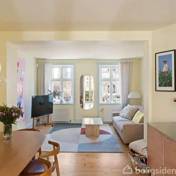 Stue med lyse vægge og trægulv, hvor en sofa står modsat et TV. Der er et spisebord med blomster på i forgrunden, omgivet af vinduer.