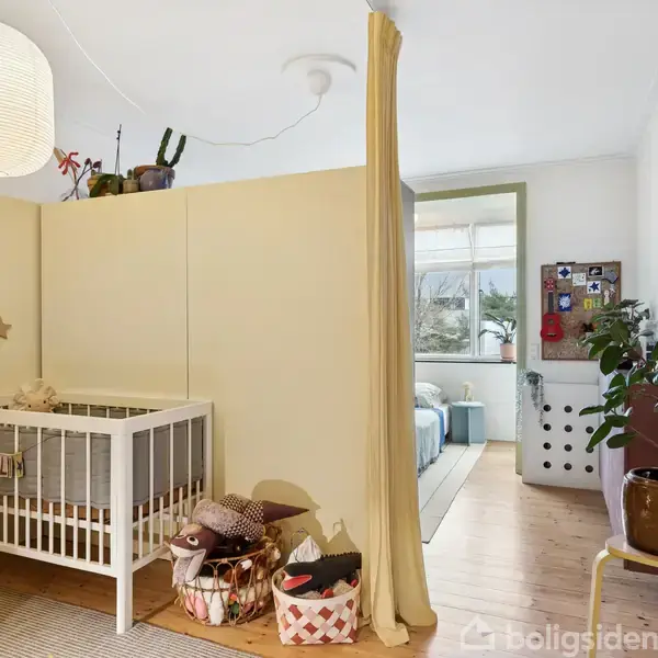 En hvid tremmeseng står ved en cremefarvet væg. Flere kurve ses på gulvet med legetøj. Rummet er opdelt med et gardin, og der er udsigt til et vindue og en plante.