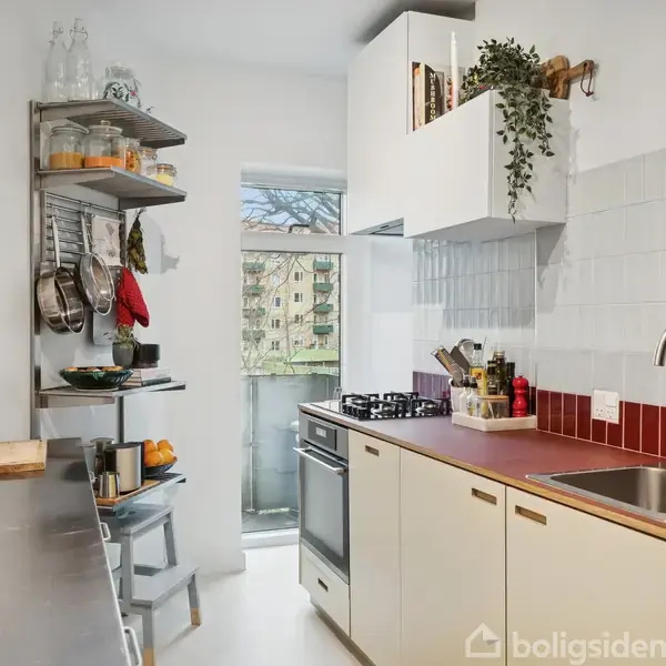 Et lille køkken med lyse, moderne skabe og rød bordplade. Komfur og vask er installeret. En hylde med glas, tallerkener og potter. Vinduet viser en udsigt over anden bygning.