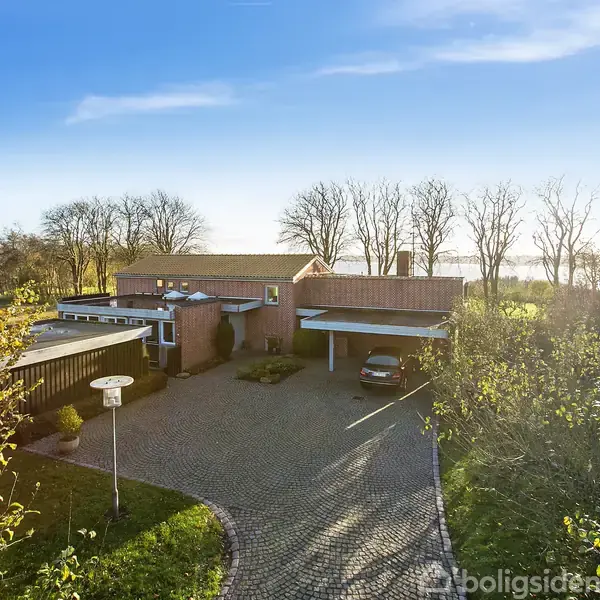 Et moderne hus i træ står stille omkranset af frodigt grønt landskab og træer. Foran er en brolagt indkørsel med en parkeret bil under en carport.