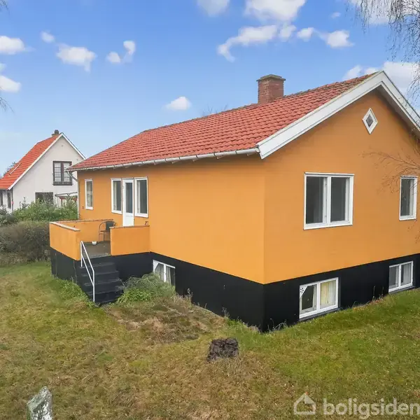 Et gult hus med rødt tegltag og hvidrammede vinduer står i en have med græs og træer. En trappe fører op til en lille terrasse. En anden hus ses i baggrunden.