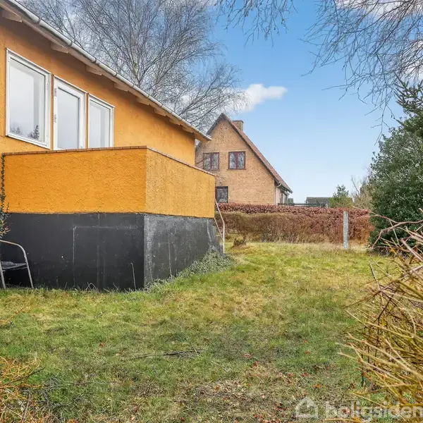 Et gult, firkantet hus med sort sokkel står stille på en velplejet græsplæne, omgivet af buske og træer, med et andet hus i baggrunden under en blå himmel.