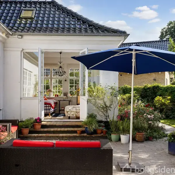 En terrasse med sort sofa og rød pude står under en blå parasol. Den åbne dør fører til en rummelig stue. Omkring er der blomsterpotter og grøn have.