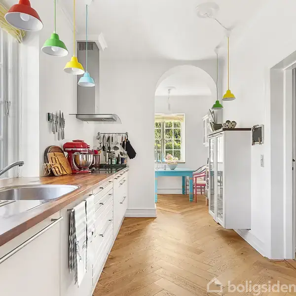 Et lyst køkken med en lang bordplade, moderne apparater og farverige lamper hængende fra loftet. I baggrunden ses en spisestue med et rundt bord og stole ved vinduet.