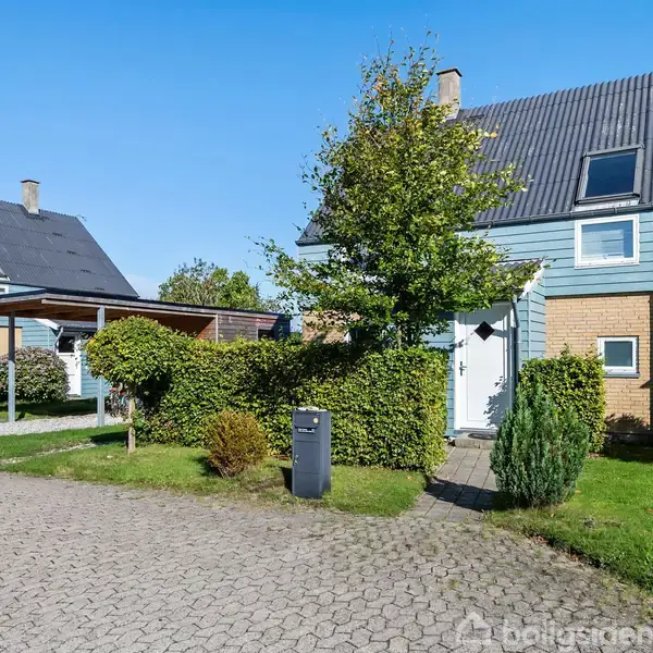 Et rækkehus med gråt tag og blå facader, omgivet af hække og grønne træer, står ved en stenbelagt vej. Parkeringspladsen er delvist dækket af en carport.