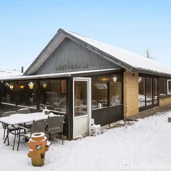 Et hus med skæve tag og vinterhave, set fra siden, beliggende i et snedækket område. Foran står et bord og stole i sneen.