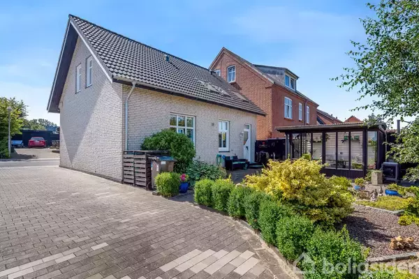 Et moderne, lyst stenhus med mørkt tag har en velholdt forhave med buske og planter. Huset ligger i et boligområde med en parkeringsplads. Skyfri, blå himmel i baggrunden.