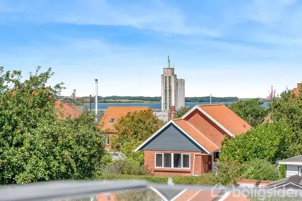 Rødt murstenshus med blåmalet gavl og rødtag, omgivet af grønne træer. I baggrunden ses en høj, moderne bygning ved en vandkant under en blå himmel.