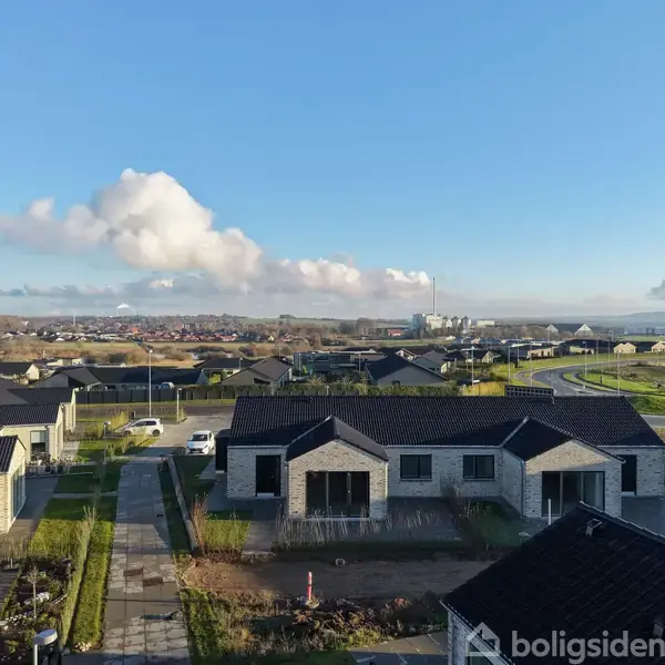 Moderne enfamiliehuse med mørke tagsten står stille i en nyligt opført forstad, omgivet af velplejede haver og åbne veje. Baggrunden viser en klar blå himmel og fjerne bakker.