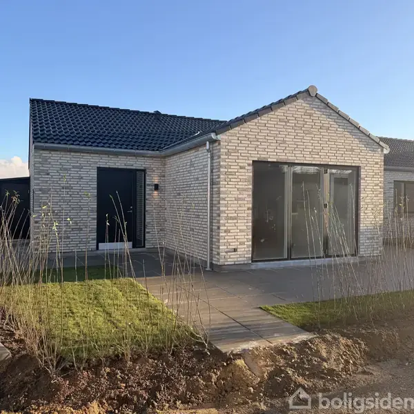 En moderne, én-etagers murstensvilla med store glasdøre, der åbner ud mod en stenbelagt terrasse, omgivet af en nyligt anlagt have under en klar blå himmel.