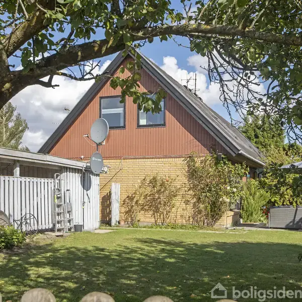 Et hus med skråt tag og to parabolantenner står i en have med grøn græsplæne, træer og en hvid hegn. Skyet himmel i baggrunden.