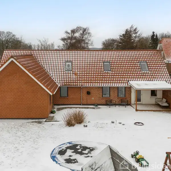 Et rødt murstenshus med skråt tag er dækket af sne. Huset har flere vinduer og et indgangsparti. I baghaven ses sneklædte buske og haveredskaber. En gynge og legetøj ligger spredt.