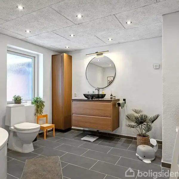 Badeværelse med rundt spejl og mørkt skab. Håndvask på skænken, badekar foran. Toilet ved vindue, planter på gulvet. Moderne, lyst interiør med grå fliser og spotlys i loftet.