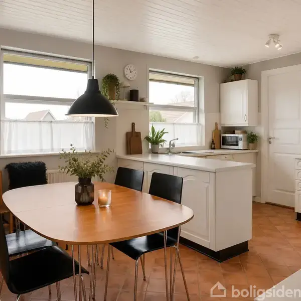 Et køkken med et træbord og sorte stole i forgrunden. En lampe hænger over bordet. Rummet har hvidt køkkeninventar, planter, og to store vinduer. Væggen har maleri og ur.
