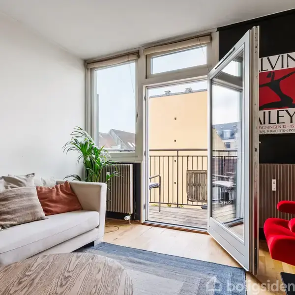 Sofa med puder står ved altandør i en lys stue med trægulv. Rød stol til højre. Plakat med teksten "MELVIN ZINEY" hænger på væggen.