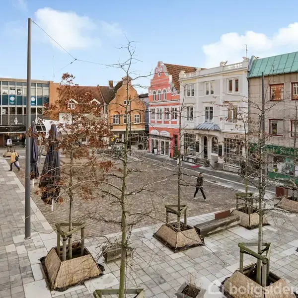 Torg omgivet af ældre bygninger i forskellige farver, hvor enkelte mennesker går over brostensbelagt plads. Omkring torvet ses træer plantet i kasser og grå fliser.