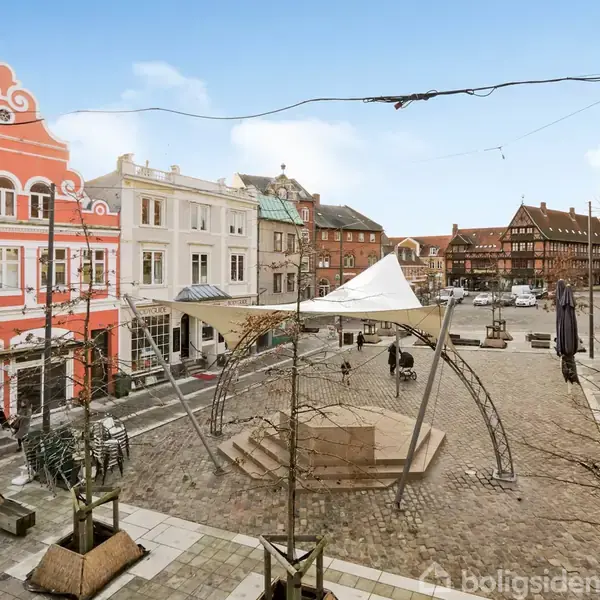 En åben plads med brosten, omgivet af farverige, gamle bygninger. Midten har en skulpturel strukturelle installation med en hvid sejldug. Træer i plantekasser står omkring pladsen.