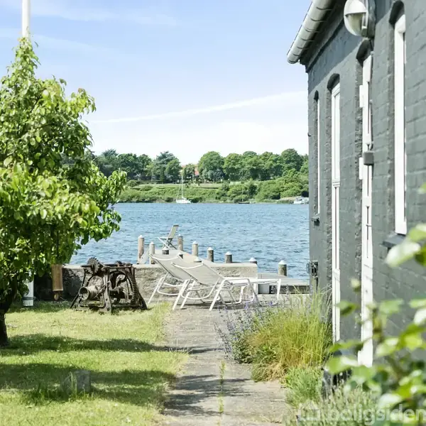 Hus ved vandet med liggestole på en mole. Træer og græsplæne omgiver området, mens blå himmel og vand ses i baggrunden.