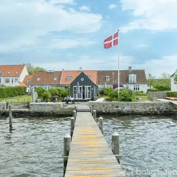 En træbro strækker sig over vandet mod et sort hus med hvidt tag. Et dansk flag vajer foran huset, omgivet af grønne haver og hvide nabohuse under en lys, overskyet himmel.