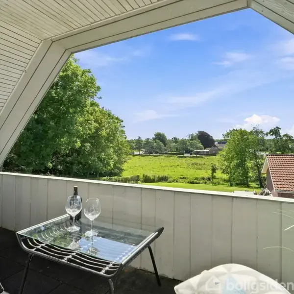 En balkon med glasbord og to stole, beliggende i en overdækket alkove. Der står to vinglas og en flaske på bordet. Udsigt til grønne marker og træer.