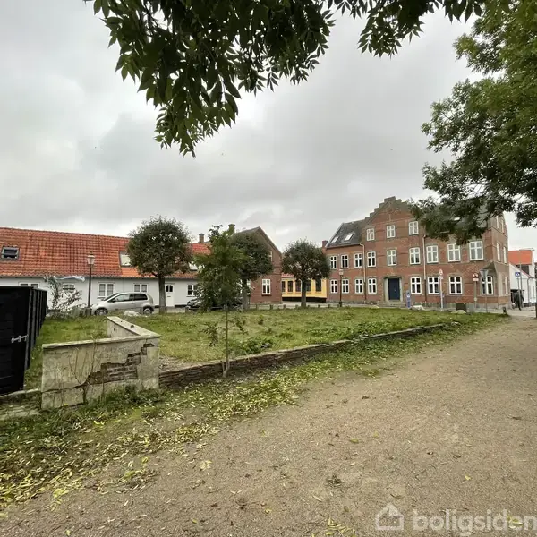 Et større, rødt murstenshus står ved siden af mindre hvide huse. Træer og buske vokser langs en grusvej i en bymæssig omgivelser.