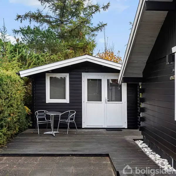 Sort træhus med hvid dør og vinduer står på en terrasse, omgivet af grønne buske og træer. To metalstole står udenfor på terrassen.