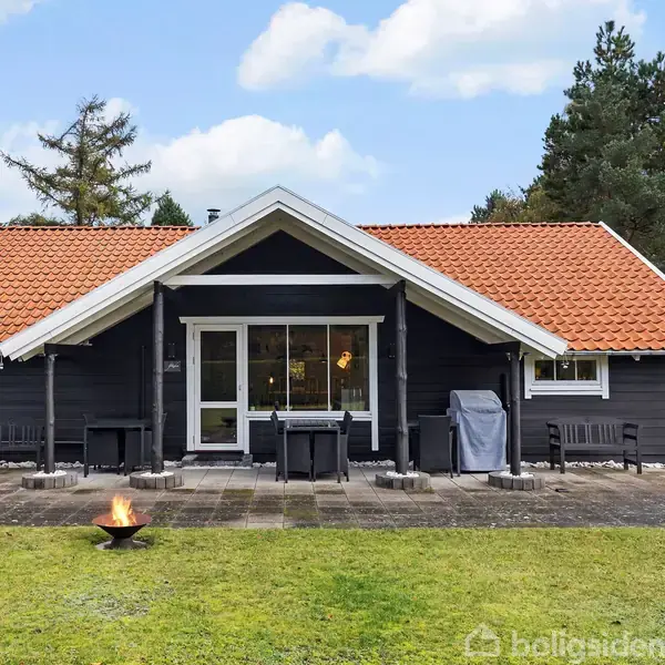 Et sort træhus med rødt tag står midt i en grøn have. Foran huset er en terrasse med møbler og en ildskål. Huset er omgivet af træer.