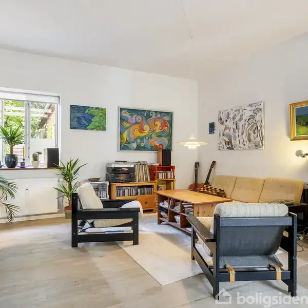 En stue med lyse farver indeholder to sofaer og et sofabord, omgivet af planter og kunstværker på væggene. En kommode og vindue bidrager til den hyggelige atmosfære.