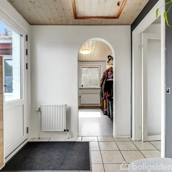 Gang med hvide vægge og lyst træloft, der leder til et andet rum gennem en bueåbning. En hvid dør med et vindue er til venstre ved siden af et træskab.