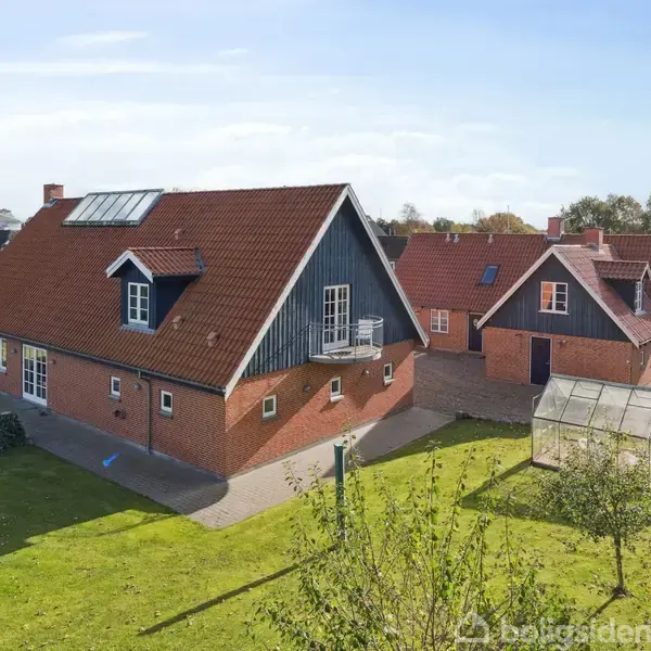 Et rødt murstenshus med rødt tegltag står stille i en grøn have omgivet af hegn. En tilhørende mindre bygning og et drivhus ses i baggrunden.