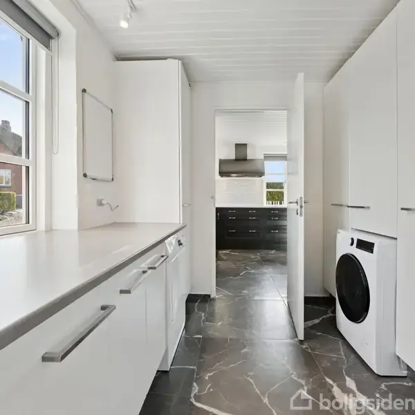 Et rummeligt bryggers med hvide skabe og en vaskemaskine til højre. Store vinduer lader lys ind, og skaber en lys atmosfære. Døren fører til et tilstødende rum.