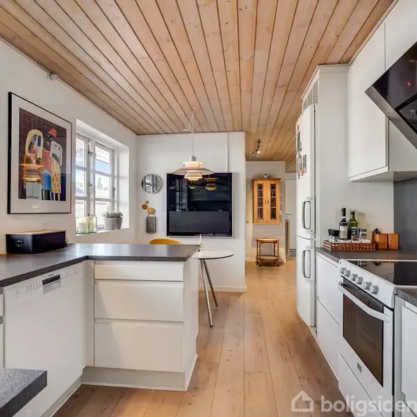 Køkken med moderne hvide skabe og stålapparater. En kogeø med barstole, træloft og vinduer giver lysindfald. Maleri og pendellampe på væggen, spiseområde i baggrunden.