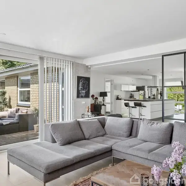 En grå sofa står midt i en stue. Store glasdøre fører ud til en terrasse. Køkkenet er i baggrunden, opdelt af en glasvæg. En vase med blomster pryder bordet.