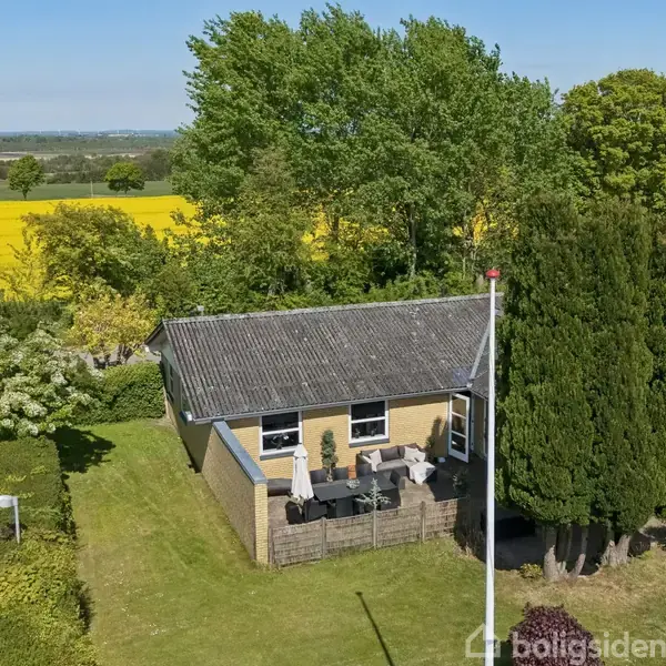 Et lille hus med terrasse står blandt træer. Huset er lavet af mursten og omgivet af en velplejet græsplæne. I baggrunden ses landbrugsmarker og en blå himmel.