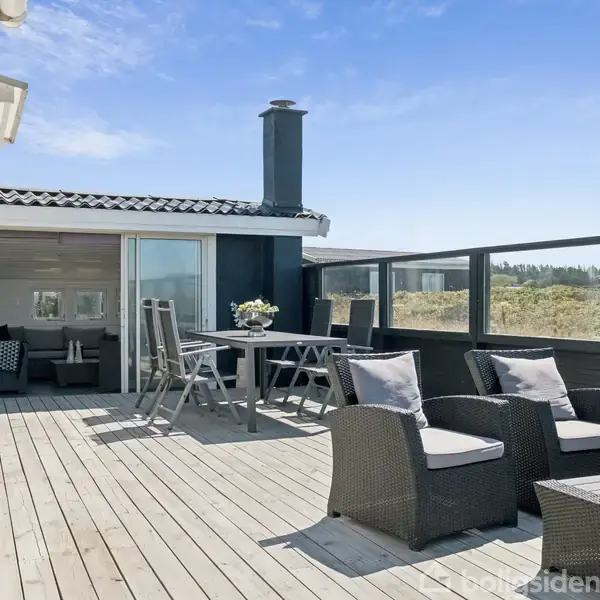 Terrassemøbler står på en rummelig træterrasse med gyngestole og et spisebord. I baggrunden ses en glasdør til stuen. Miljøet er solrigt med udsigt over naturen.