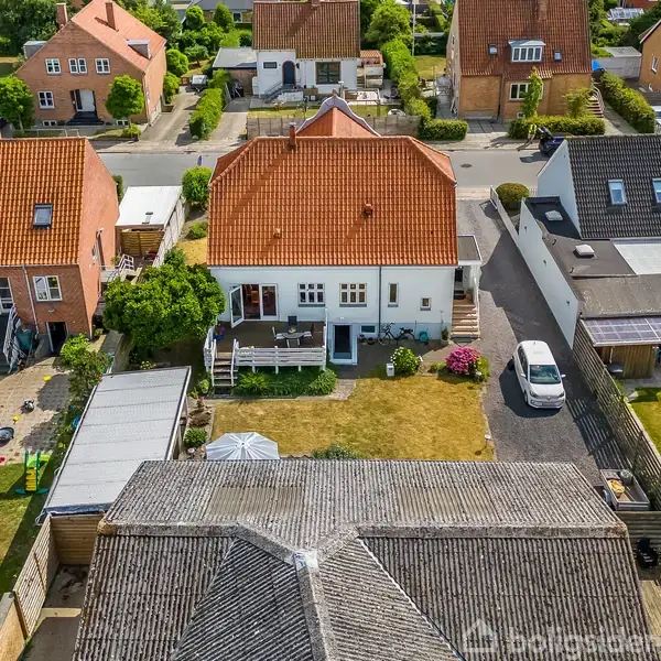 Hvidt hus med rødt tegltag står stille i en forstadshave. På terrassen er der nogle havemøbler. Haven er omkranset af andre huse med velplejede græsplæner og blomsterbede.