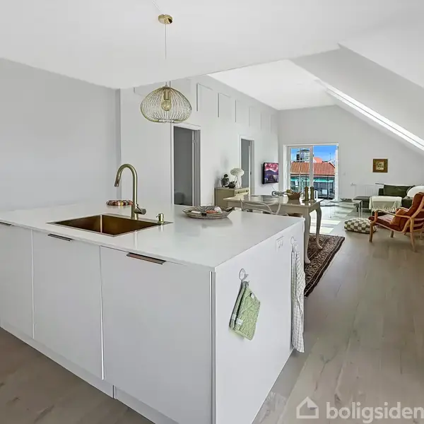 Køkkenø med indbygget vask i en lys, åben stue. En sofa og lænestol er opstillet med et tæppe på trægulv, mens en dør fører til en balkon.