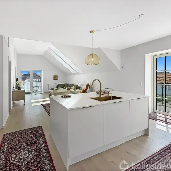 Køkkenø med vask og pendellampe i en lys og rummelig stue. Rummet har et skråt loft, tæpper på gulvet, og franske døre der åbner mod en balkon med udsigt.