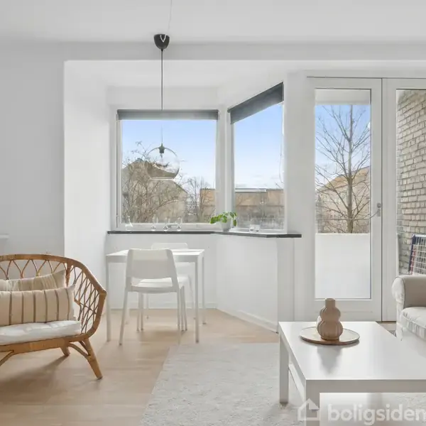 En stue med en sofa og bord, der har en keramikdekoration, ved siden af et vindue. Der er også en lænestol og et lille spisebord med to stole foran vinduet.