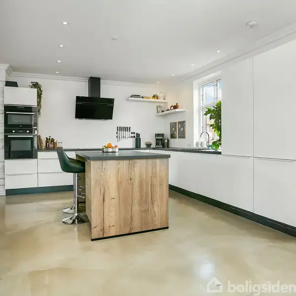 En lys stue med en grå hjørnesofa står langs vinduerne. En sort sofabord står foran sofaen. I baggrunden ses et spisebord med stole og et stort maleri på væggen. En plante står ved siden af en skab.