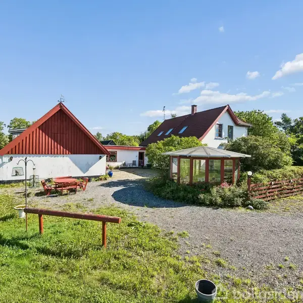Rødt og hvidt hus med spidstag står ved siden af en rød lade. Hagen er omgivet af frodige træer og en grusvej. Sollys belyser de grønne omgivelser.