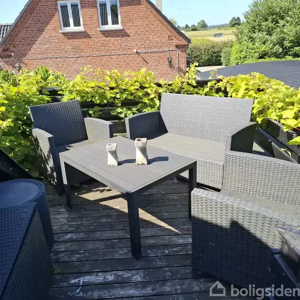 Terrasseområde med grå kurvemøbler arrangeret omkring et firkantet bord. To små dekorationer står på bordet. Røde murstensbygninger og grønne buske omgiver området i solskin.